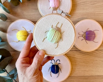 May include: Five embroidered hoop art pieces featuring colorful beetles with gold accents. The hoops are displayed on a wooden surface.