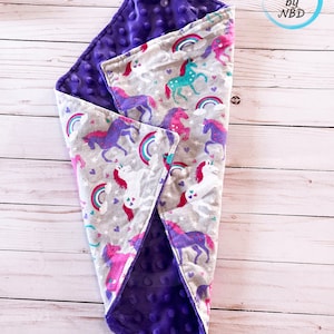 Puede incluir: Una manta para bebé de color morado y gris con estampado de unicornio y arcoíris. La manta es suave y acogedora, perfecta para un bebé o un niño pequeño.