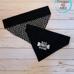 Può includere: Un bandana per cani nero e bianco con un bordo nero e una stampa di ossa bianche. Il bandana ha una sovrapposizione nera con il testo "BAD to the BONE" in bianco con una grafica di ossa bianca.