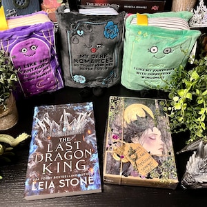 May include: An assortment of fantasy-themed items, including three plush book covers with embroidered quotes and whimsical faces. Two books are visible, one titled "The Last Dragon King" and another with a woman's portrait. Decorative greenery and dragon figurines complete the scene.