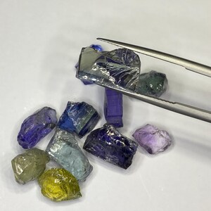 May include: A collection of raw gemstones in various colors, including blue, purple, green, and yellow. A pair of tweezers holds one of the gemstones. The gemstones are rough-cut and have a natural, unpolished appearance.