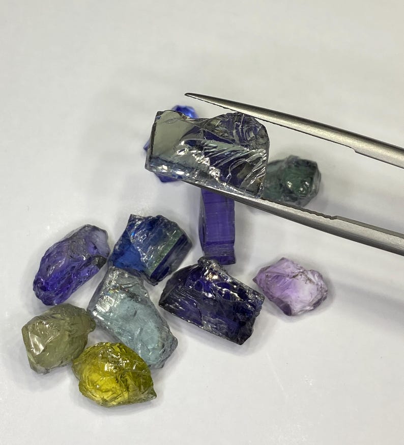 May include: A collection of raw gemstones in various colors, including blue, purple, green, and yellow. A pair of tweezers holds one of the gemstones. The gemstones are rough-cut and have a natural, unpolished appearance.