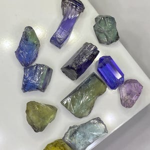 May include: A collection of raw gemstones in various colors, including blue, green, purple, and yellow. The gemstones are irregularly shaped and appear to be uncut, with a rough, natural texture. The arrangement of the stones is scattered across a white surface.