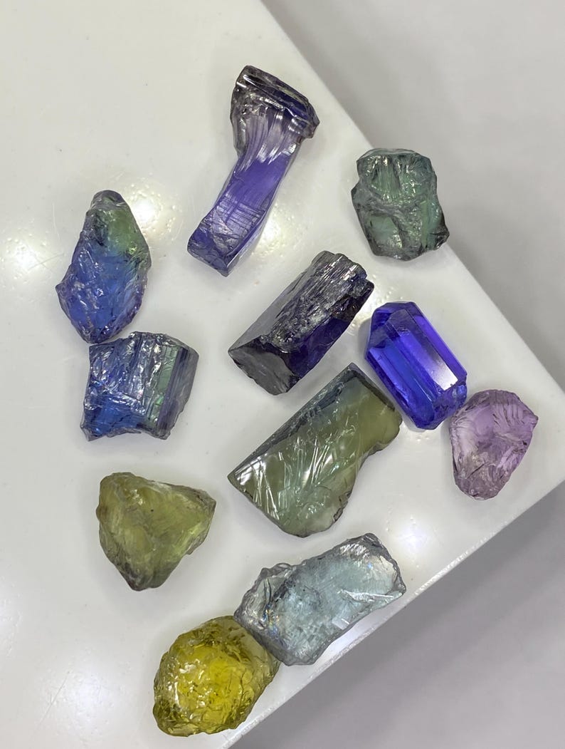 May include: A collection of raw gemstones in various colors, including blue, green, purple, and yellow. The gemstones are irregularly shaped and appear to be uncut, with a rough, natural texture. The arrangement of the stones is scattered across a white surface.