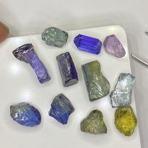 May include: A collection of raw gemstones in various colors, including blue, purple, green, and yellow. The gemstones are displayed on a white surface, showcasing their natural textures and shapes. Some are rectangular, others are more organic.