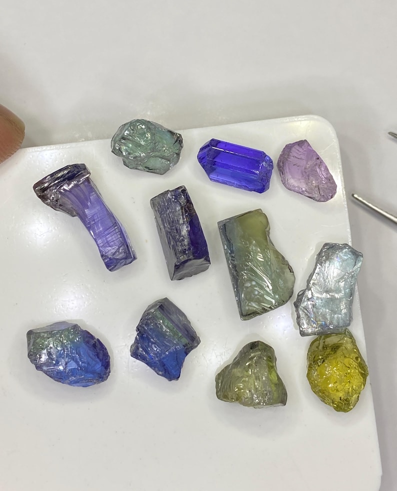 May include: A collection of raw gemstones in various colors, including blue, purple, green, and yellow. The gemstones are displayed on a white surface, showcasing their natural textures and shapes. Some are rectangular, others are more organic.