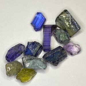 May include: A collection of raw gemstones in various colors, including blue, purple, green, and yellow. The gemstones are irregularly shaped and appear to be uncut, with a crystalline structure. The background is a plain white surface.