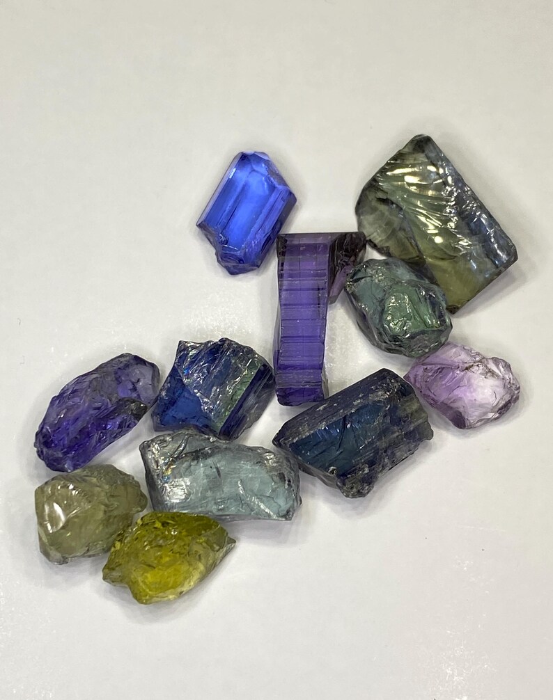 May include: A collection of raw gemstones in various colors, including blue, purple, green, and yellow. The gemstones are irregularly shaped and appear to be uncut, with a crystalline structure. The background is a plain white surface.