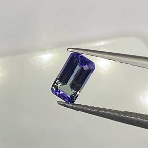 Könnte beinhalten: Ein rechteckiger, facettierter Tansanit-Edelstein, der von einer Pinzette gehalten wird. Der Edelstein zeigt einen tiefviolett-blauen Farbton mit grünen Nuancen. Die Facetten fangen das Licht ein und zeigen die Klarheit und Brillanz des Steins. Der Hintergrund ist ein weiches, neutrales Weiß.