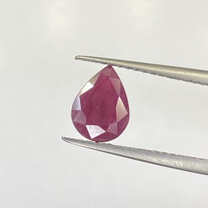 以下が含まれることがあります： 銀色のピンセットで保持された、ファセット加工された洋梨型のルビー。ルビーは、光を捉える複数のファセットを持つ深い赤色を示しています。宝石は、その色と形を際立たせるために、無地の白い背景に配置されています。