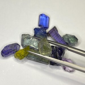May include: A collection of raw gemstones in various colors, including blue, green, purple, and yellow. The gemstones are held by a pair of silver tweezers. The gems are irregularly shaped and appear to be uncut.