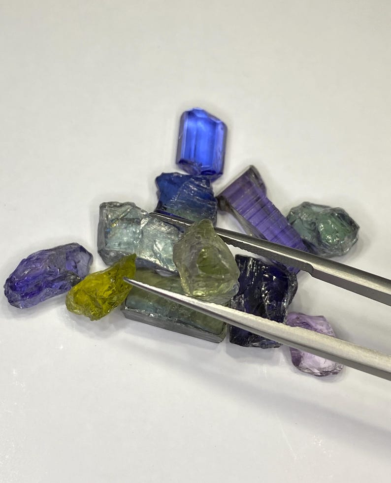 May include: A collection of raw gemstones in various colors, including blue, green, purple, and yellow. The gemstones are held by a pair of silver tweezers. The gems are irregularly shaped and appear to be uncut.