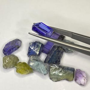 May include: A collection of raw gemstones in various colors, including blue, green, and purple. A pair of tweezers holds a vibrant blue gemstone. The gemstones are rough-cut and appear to be natural, with visible textures and imperfections.