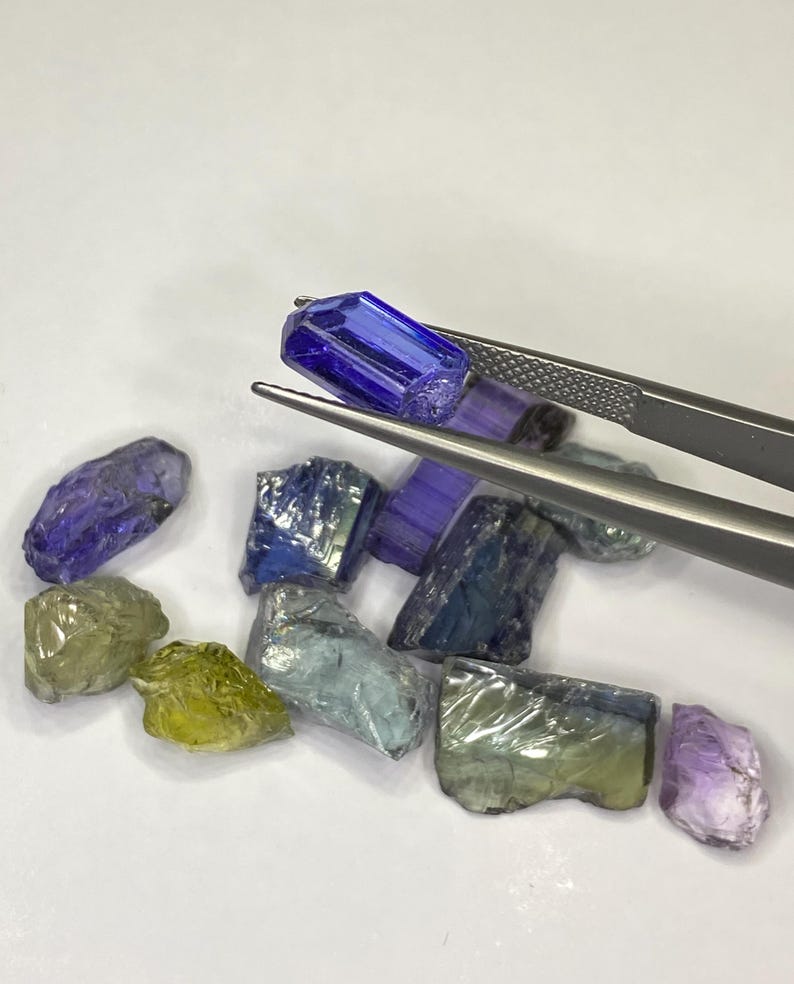 May include: A collection of raw gemstones in various colors, including blue, green, and purple. A pair of tweezers holds a vibrant blue gemstone. The gemstones are rough-cut and appear to be natural, with visible textures and imperfections.