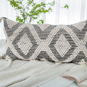 以下が含まれることがあります： 白黒の幾何学模様の長方形の装飾用枕。 枕は、小さな隆起したドットと黒いストライプを備えたテクスチャ加工された表面を持っています。 各端にはタッセルが付いています。 枕は白い表面にあります。