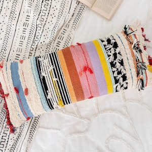 Peut inclure: Un long coussin rectangulaire avec un motif rayé coloré. Le coussin est fait d'un tissu tissé avec une bordure en franges. Les couleurs des rayures incluent le rouge, l'orange, le jaune, le vert, le bleu et le violet.