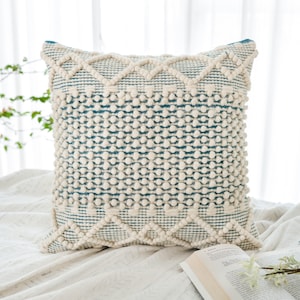 Op de afbeelding: Een vierkant kussen met een geometrisch patroon in wit en blauw. Het kussen heeft een textuur met kleine witte pompoentjes rond de randen.