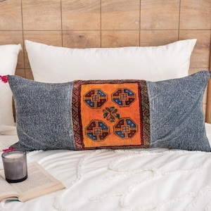 Op de afbeelding: Een rechthoekig sierkussen met een oranje middenpaneel met een geometrisch ontwerp in blauw, rood en bruin. De uiteinden van het kussen zijn van een grijze stof met textuur en hebben aan elk uiteinde kwastjes. Het kussen ligt op een wit bed.