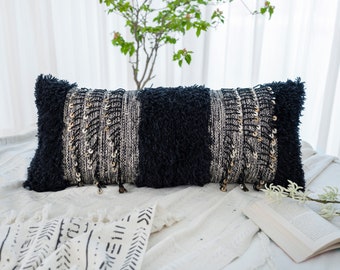 May include: Black and white patterned lumbar pillow with gold accents and a fluffy texture. The pillow is on a white bedspread with a black and white patterned throw blanket.
