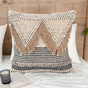 Può includere: Un cuscino decorativo con un motivo geometrico in tonalità beige, marrone e grigio. Il cuscino ha un bordo a frangia e una superficie testurizzata.