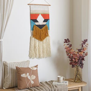 May include: A colorful woven wall hanging with geometric patterns in red, blue, black, and white, accented by a mustard-colored fringe. The wall hanging is suspended from a wooden dowel. Decorative pillows and a vase of flowers are also in the image.