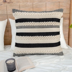 May include: A black and white striped throw pillow with a textured woven design. The pillow is on a white bed with a wooden headboard.