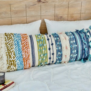 May include: A long, decorative cushion with a white background and colourful stripes in shades of blue, green, orange, and brown. The cushion has a fringe trim and is decorated with small, round pom-poms.