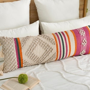 Könnte beinhalten: Ein langes, rechteckiges Kissen mit einem gestreiften Muster in Beige, Creme, Orange, Rosa und Grün. Das Kissen hat eine strukturierte Oberfläche und Quasten an den Enden.