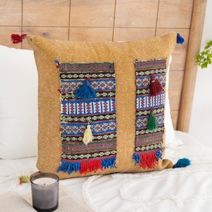 May include: Mustard-coloured cushion with embroidered panels featuring geometric patterns in blue, red, and black. The cushion has colourful tassels and is on a white bed with a candle and book.