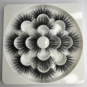 Puede incluir: Una bandeja circular blanca con diez compartimentos individuales, cada uno de los cuales contiene un juego de pestañas postizas. Las pestañas son negras y parecen estar hechas de fibras sintéticas.