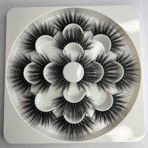 Puede incluir: Un conjunto de siete pestañas postizas negras dispuestas en una bandeja circular blanca. Las pestañas son gruesas y voluminosas, con una apariencia plumosa. La bandeja tiene compartimentos individuales para cada par de pestañas.