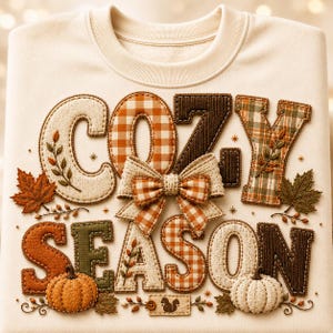 Puede incluir: Una sudadera color crema con el texto COZY SEASON en letras grandes, texturizadas y con varios patrones. El diseño incluye un lazo central de cuadros vichy, calabazas naranjas y blancas, hojas de otoño y un pequeño parche rectangular con una ardilla.