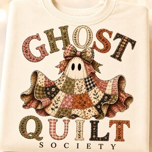 Pode incluir: Uma camisola de cor creme com um gráfico de um fantasma com um laço, rodeado pelo texto GHOST QUILT SOCIETY. O fantasma é representado com um padrão de colcha de retalhos em várias cores suaves, incluindo rosa, verde e castanho.