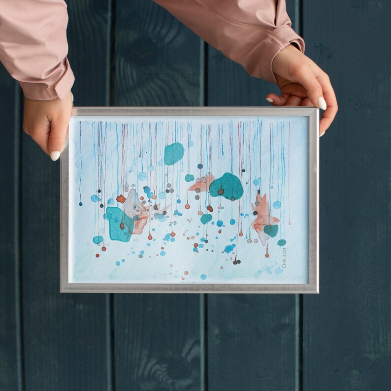 My handmade unique abstract mixed media work called Rain. Blue, metallized green and orange shades like rain drops. Placed in a mockup frame above a dark green background and held by a woman wearing a light pink jacket.