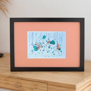 My handmade unique abstract mixed media work called Rain. Blue, metallized green and orange shades like rain drops. Placed in a black mockup frame with an orange mount, on a wooden table.