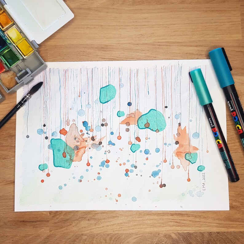 My handmade unique abstract mixed media work called Rain. Blue, metallized green and orange shades like rain drops. Placed on a wooden table with drawing and painting tools around it.