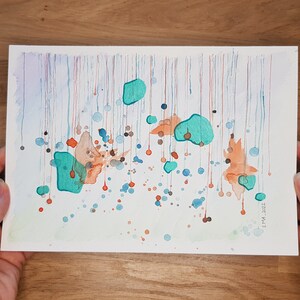 My handmade unique abstract mixed media work called Rain, held in my hands. Blue, metallized green and orange shades like rain drops. Placed on a wooden table.