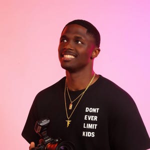 May include: A black t-shirt with the white text "DONT EVER LIMIT KIDS" is worn by a person holding a black camera with a flash. Gold necklaces and a black watch are also visible. The background is a pink and purple gradient.