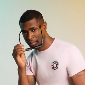 Puede incluir: Camiseta rosa claro con un diseño gráfico negro y rojo de una cámara y la letra "D" en el pecho. La persona sostiene gafas con montura negra. El fondo tiene un degradado de turquesa y amarillo.