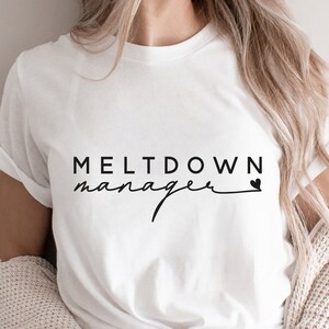 May include: White t-shirt with black text that reads "MELTDOWN manager" with a small heart symbol.