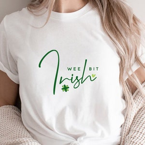 Könnte beinhalten: Weißes T-Shirt mit einem grünen Kleeblatt und dem Text "Wee Bit Irish" in einer kursivschrift.