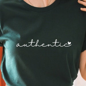 May include: A dark green t-shirt with the word "authentic" written in white cursive with a small white heart after the word. The person wearing the shirt is standing in front of a rack of clothes.