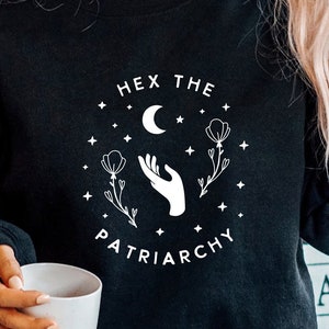 May include: Black sweatshirt with a white graphic design that says "Hex the Patriarchy" with a crescent moon, stars, and a hand reaching for a flower.