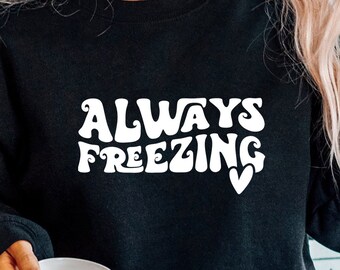 以下が含まれることがあります： 白い文字で「ALWAYS FREEZING」と書いてある黒いスウェットシャツ。小さなハートのマークが付いている。