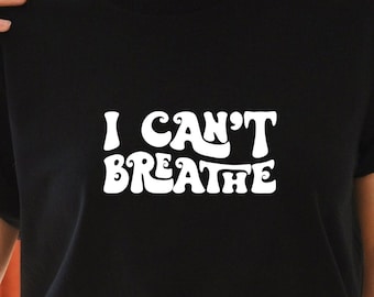 May include: Black t-shirt with white text that reads "I CAN'T BREATHE" in a retro font. The shirt is being worn by a person with curly hair and a brown belt.