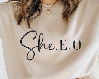 Puede incluir: Una sudadera beige con el texto "She.E.O" en letras de escritura negras. La sudadera la lleva puesta una persona que lleva pantalones marrones.