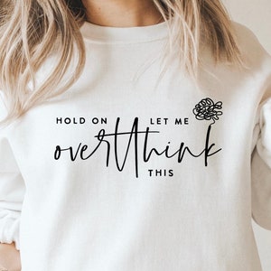 May include: White sweatshirt with the text "HOLD ON LET ME overthink THIS" in black script. A small black line drawing of a tangled brain is above the word "THIS". The sweatshirt is paired with light blue jeans.