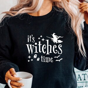 Puede incluir: Sudadera negra con texto blanco que dice "it's witches time" con una bruja volando en una escoba y murciélagos y estrellas alrededor del texto.