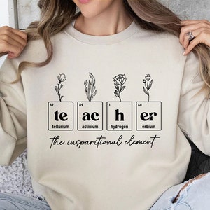 May include: A beige sweatshirt with a graphic design of the word "teacher" spelled out using chemical element symbols. The graphic also includes small floral illustrations. The text below the graphic reads "the inspirational element."