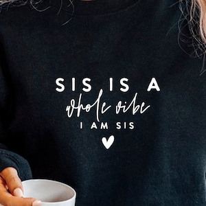 May include: Black sweatshirt with white text that reads "Sis is a whole vibe I am sis" with a small heart symbol.